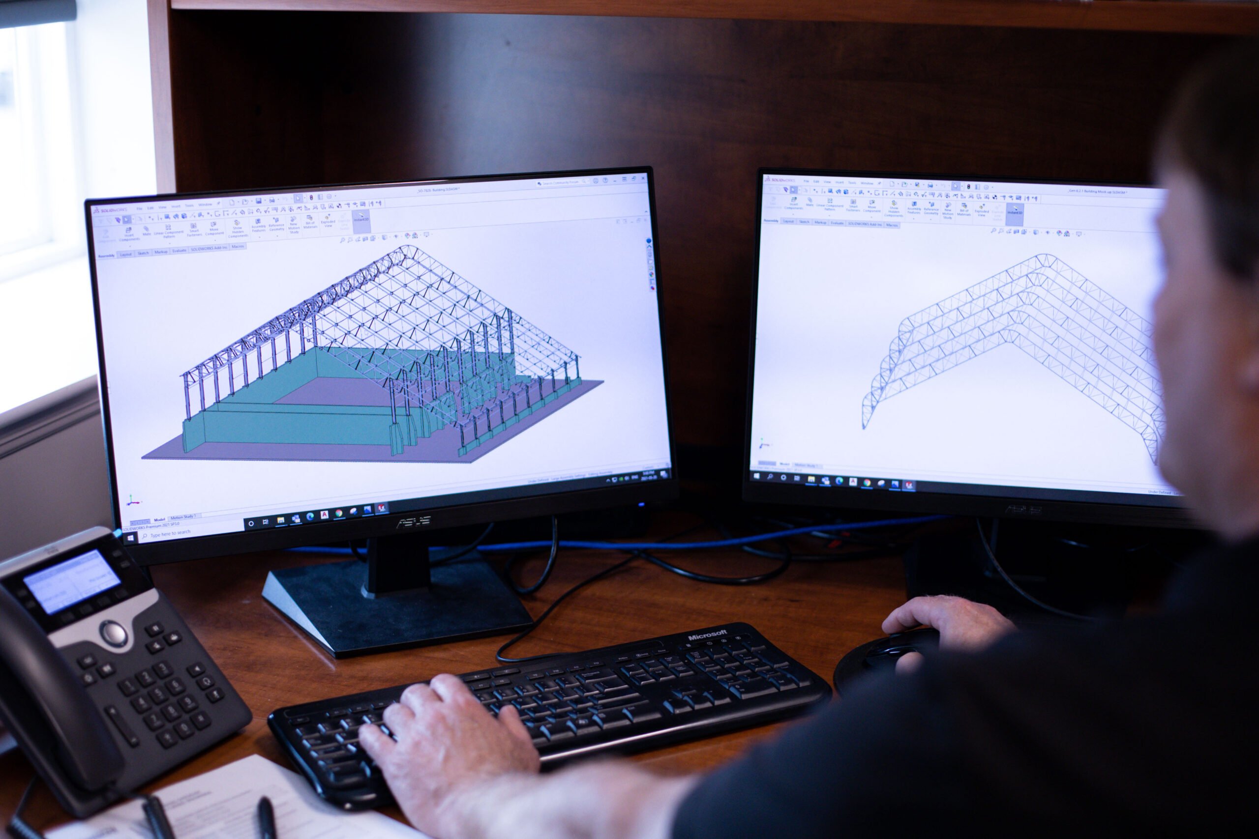 An engineer's hands on a keyboard and mouse, working on dual computer monitors displaying 3D CAD designs of custom engineered fabric structures, demonstrating the detailed engineering process for unique projects.