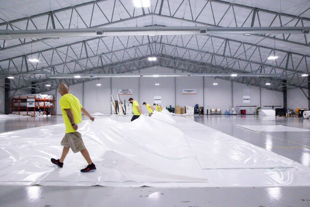 Workers in a large, well-lit manufacturing facility carefully unrolling and positioning a massive white fabric sheet, illustrating the meticulous assembly process for custom engineered fabric structures. Various rolls of fabric and industrial equipment are visible in the background, emphasizing the scale and precision involved in tailored fabric building custom design.