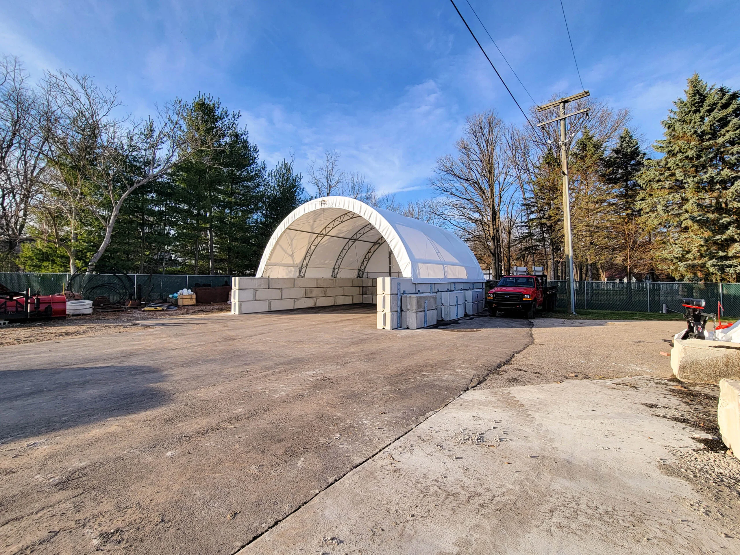 Precast Concrete Block Fabric Building Foundations - Britespan A large white fabric building with a robust foundation of stacked precast concrete blocks, situated on an industrial lot. A red pickup truck is parked next to the structure, with trees and a utility pole visible under a clear blue sky, highlighting a practical fabric building foundation solution.