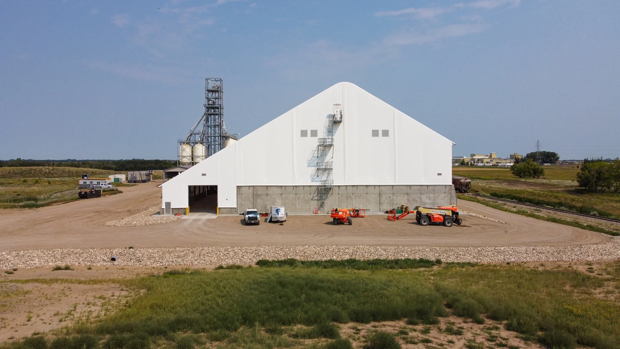 Commercial Fabric Building: Navigating Permit Requirements - Britespan Large white fabric building at a commercial salt storage facility, with a tall grain silo, rail tracks, and construction equipment, illustrating the type of structure that requires careful navigation of fabric building permits.