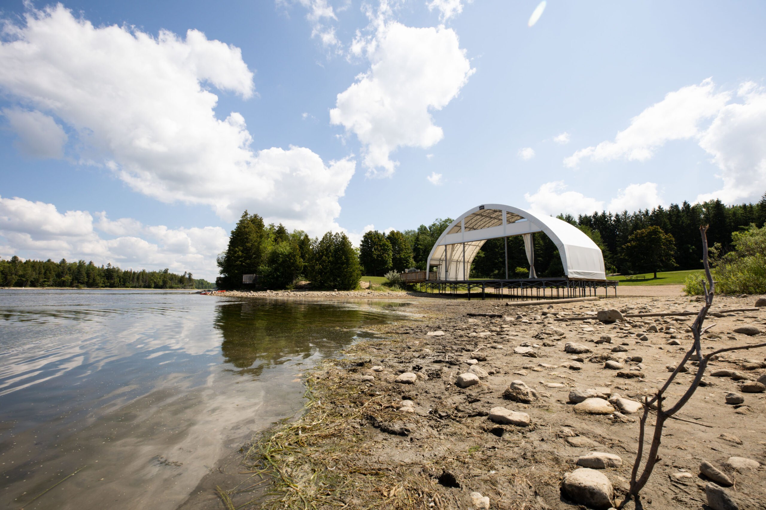 A large white relocatable fabric structure, ideal for flexible temporary building solutions, stands on a raised platform beside a muddy lake shoreline with lush green trees in the background under a bright blue sky with scattered clouds.