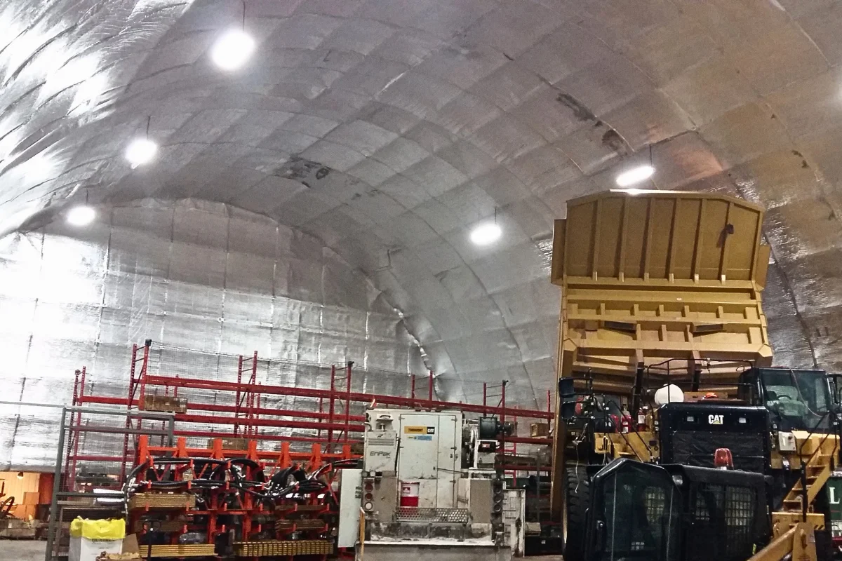 Interior view of a large fabric structure showcasing its silver insulated ceiling and walls, illuminated by bright lights. Industrial equipment, including a large yellow machinery and storage racks, are visible inside, demonstrating effective fabric structure insulation.
