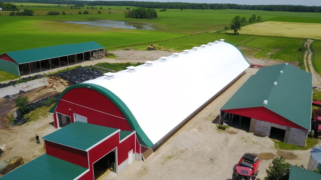 Fabric Covered Equipment Storage for Dairy Farm Expansion - Britespan Aerial view of a large white fabric covered equipment storage building with a curved roof attached to a red dairy barn, surrounded by green fields.