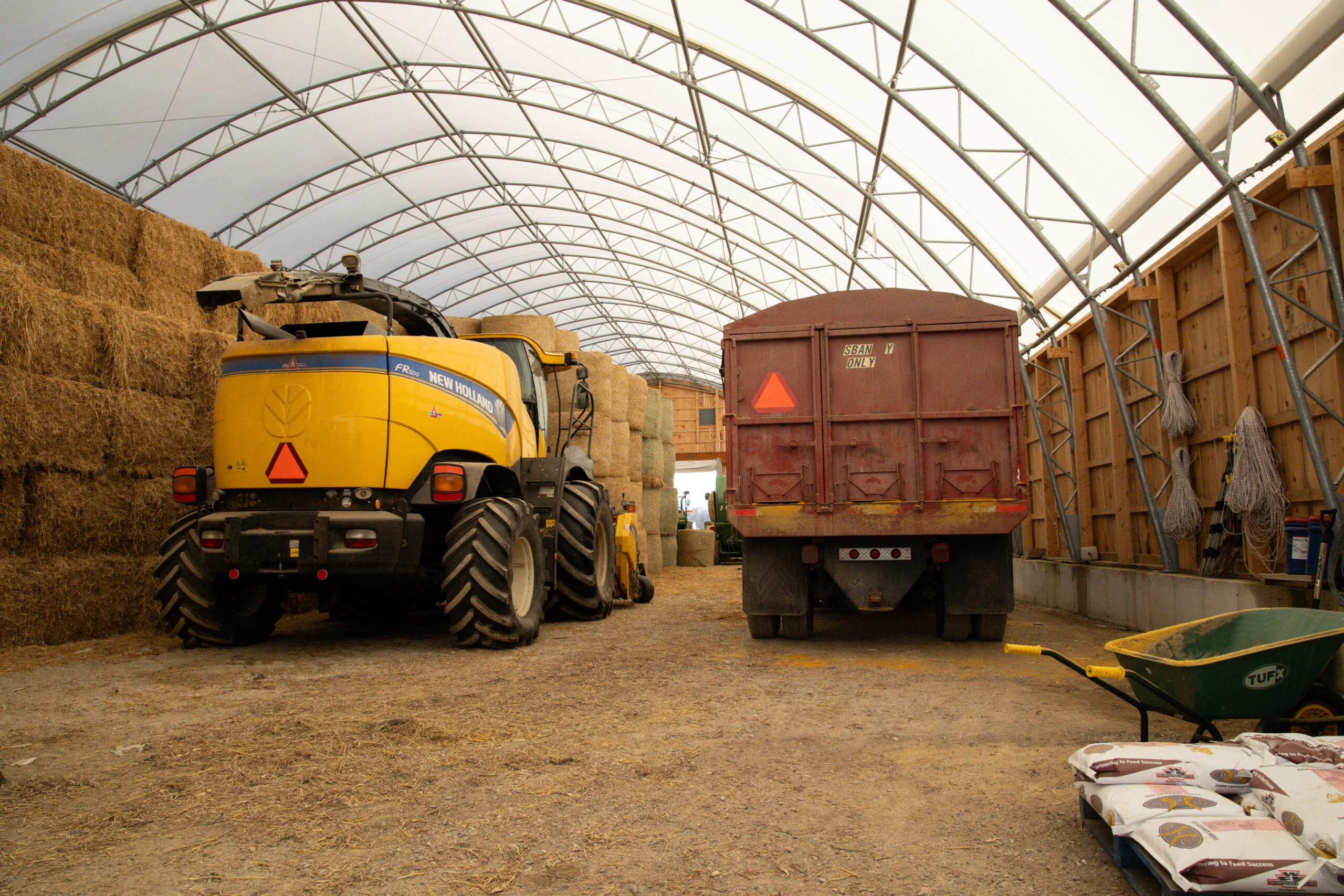 Farm equipment storage inside a clear span fabric building with hay bales, loader, and trailer in a bright, open layout