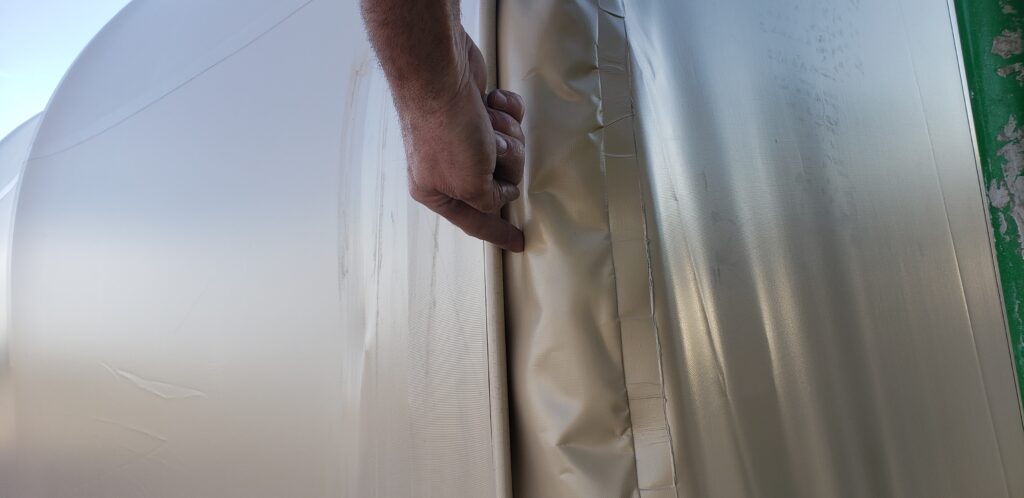 Image of the exterior of a fabric building end wall truss with hand pointing to loose fabric ready for more tensioning with sun shining. Spring Fabric Building Maintenance